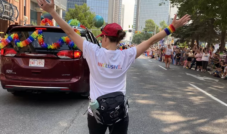 Individual with arms raised in the shape of a y with a white t shirt on that says inclusive on the back