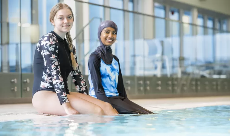 Two individuals in swimming gear sit on the ledge of an indoor pool with smiles on their faces.